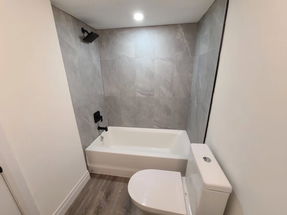 Tub alcove with large grey marble-look wall tile, matte black shower trim and wood-look floor