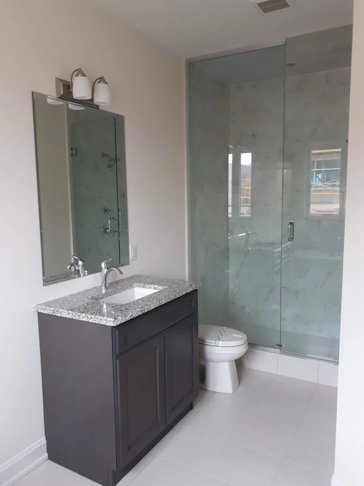 Bathroom with dark grey shaker vanity, granite top, frameless glass shower and white floor tile