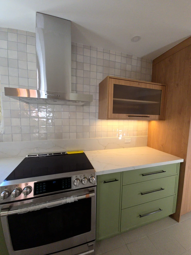 Sage green cabinets with stainless range, chimney hood, white zellige-style tile and wood shelving