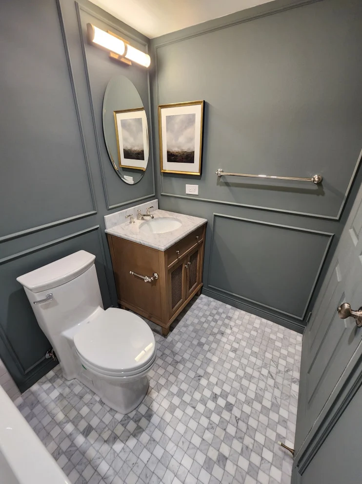 Sage grey panelled bathroom with wood vanity, oval mirror, toilet and marble basketweave floor