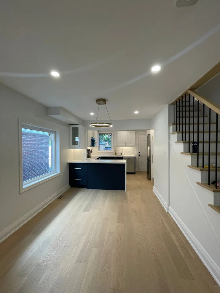 Open main floor with light oak floors, navy kitchen peninsula, ring pendant and black stair railing