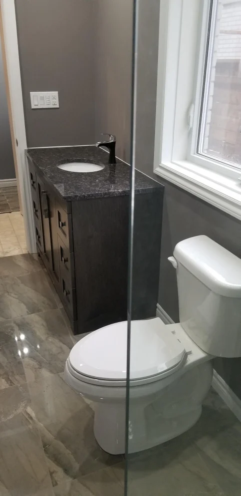 Dark vanity with granite top and white toilet beside a window on glossy tile floor