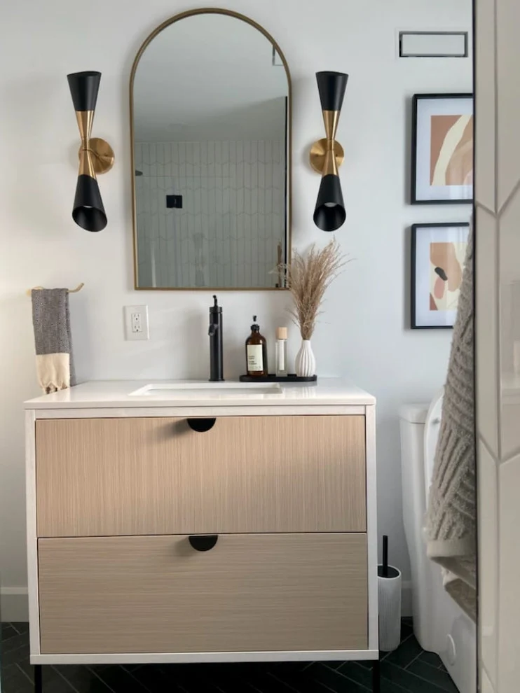 Fluted light wood vanity with white top, arched gold mirror and black and brass sconces