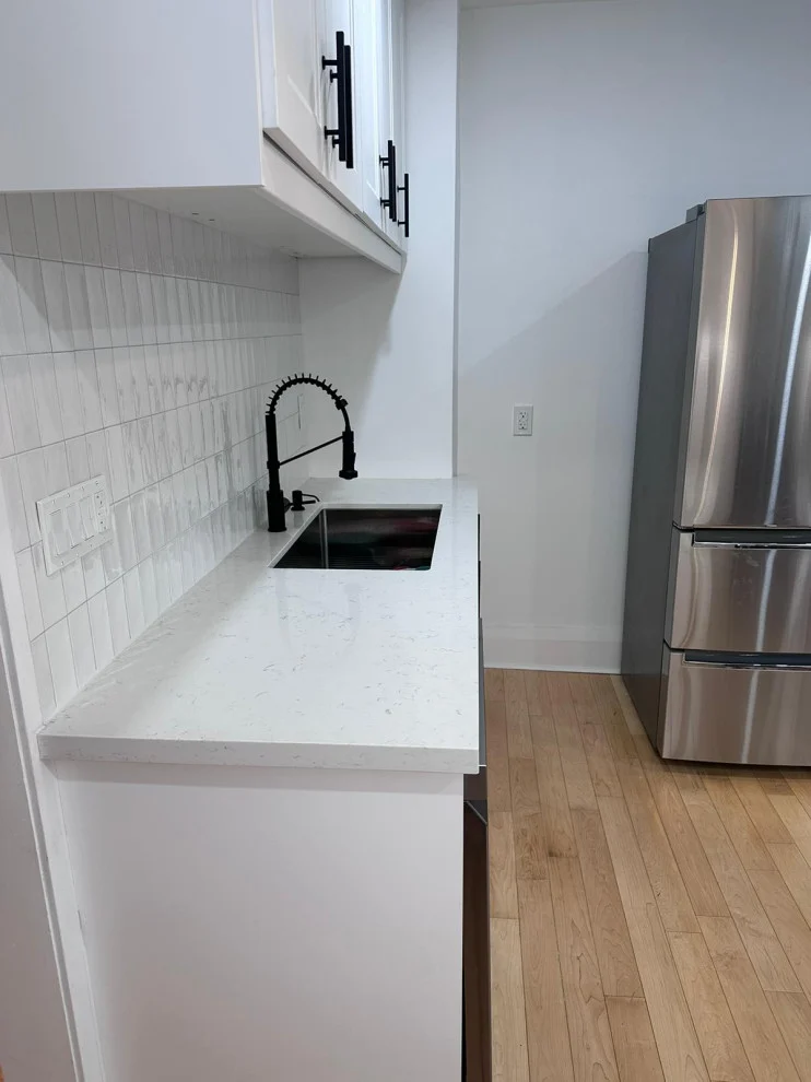 White quartz counter with black faucet, stainless fridge and light hardwood flooring