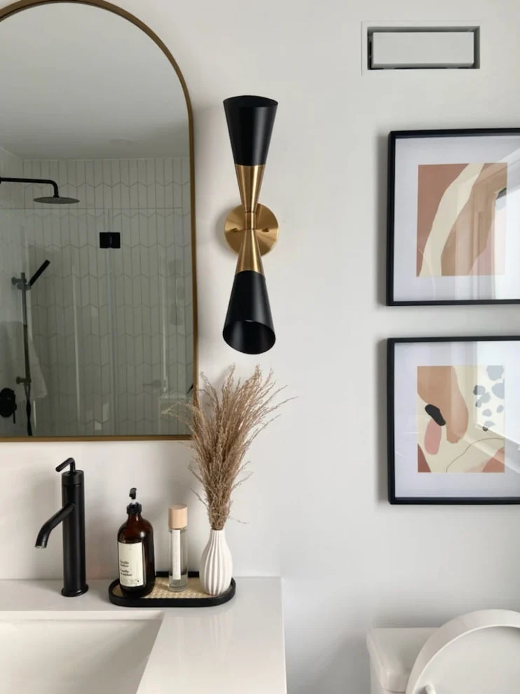 Black faucet on white counter beneath arched gold mirror with cone sconce and framed art