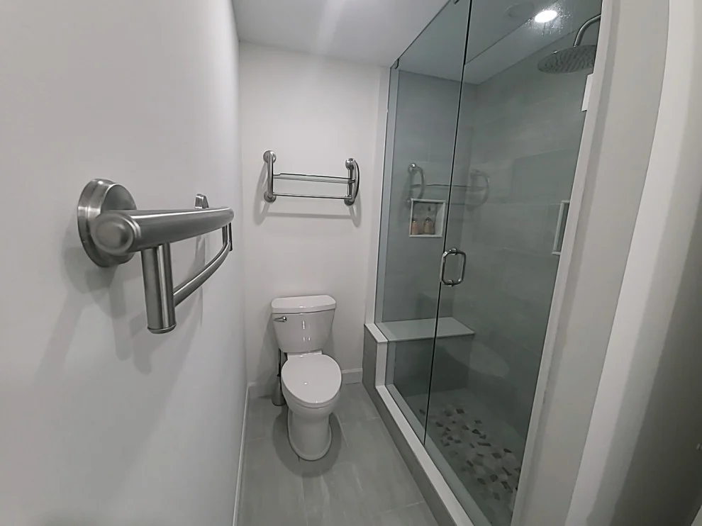 Bathroom with grab bar, towel rack, toilet and glass shower with grey tile and pebble floor