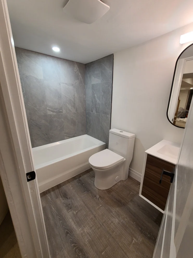 Small bathroom with tiled tub, one-piece toilet, walnut floating vanity and black framed mirror