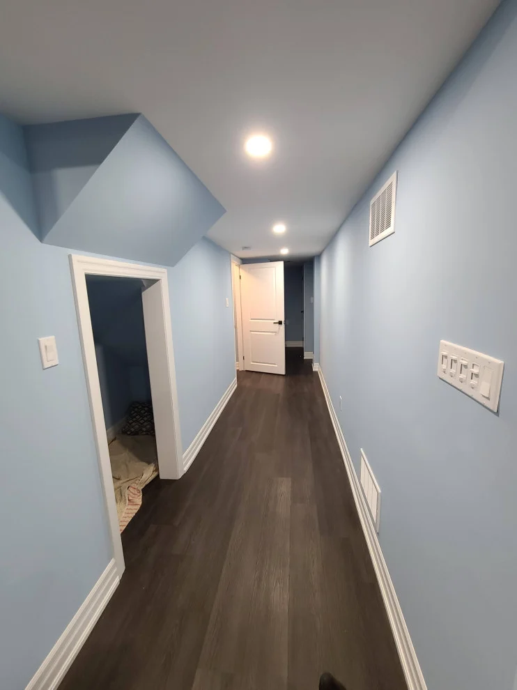 Basement hallway with light blue walls, white trim, pot lights and dark vinyl plank floor