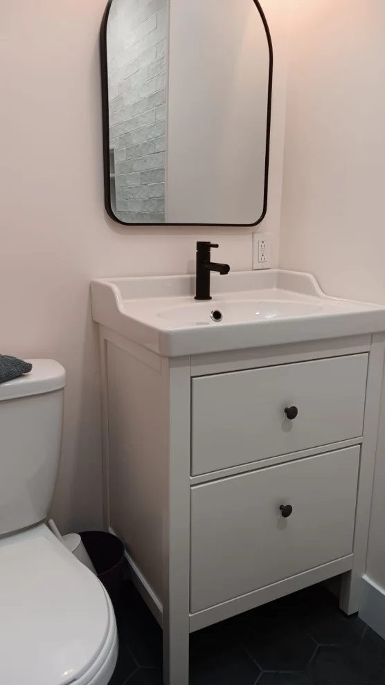 White vanity with matte black faucet below arched black framed mirror on charcoal hex tile