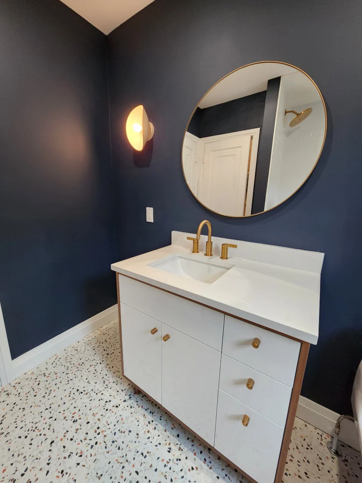 Navy blue bathroom wall with round brass mirror, white vanity with gold hardware and terrazzo floor