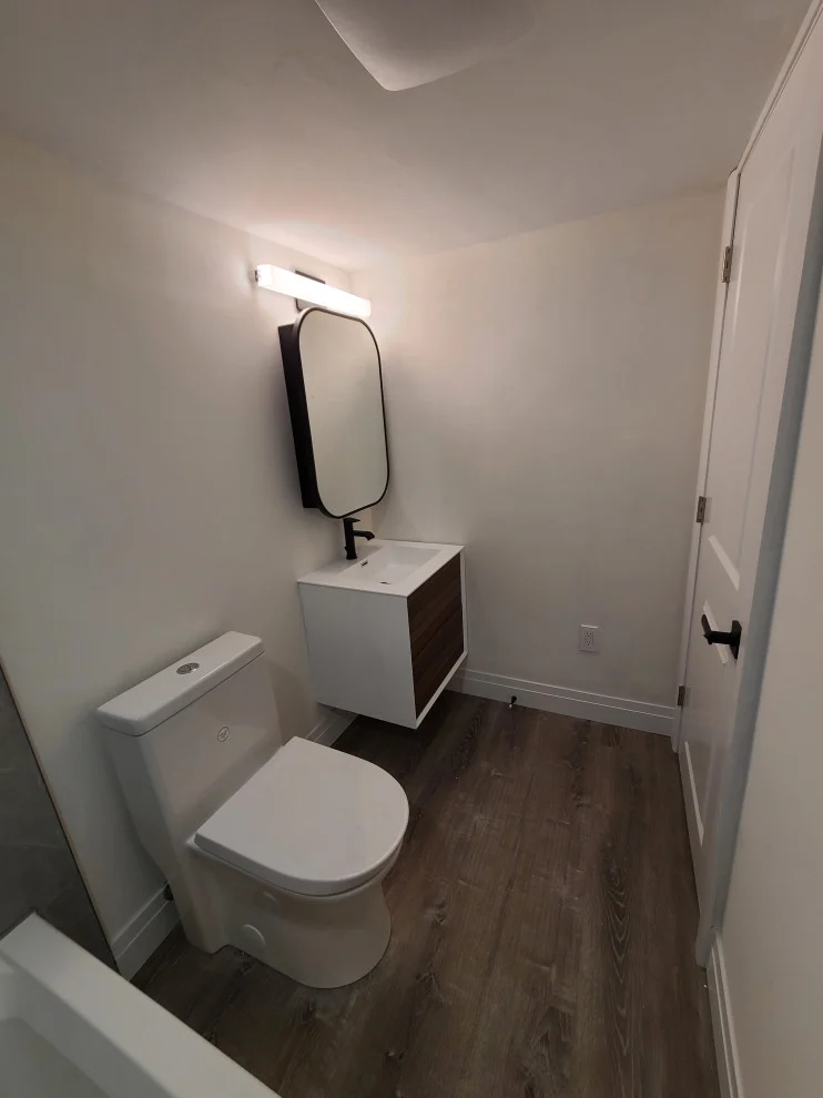 Floating walnut and white vanity with black rounded mirror, toilet and grey vinyl plank floor