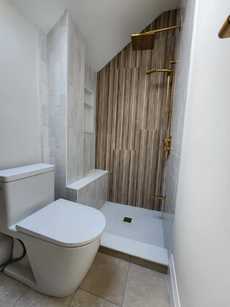 Toilet beside walk-in shower with wood-grain accent wall, white tile and gold fixtures