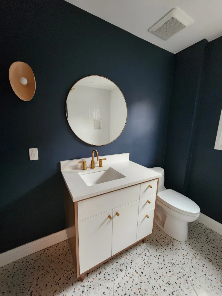 Wide view of navy bathroom with white vanity, round mirror, gold sconce, toilet and speckled floor
