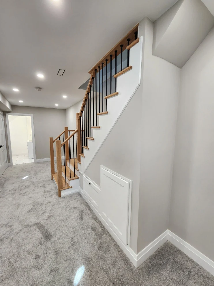 Finished basement with grey carpet, pot lights and open staircase with oak railing