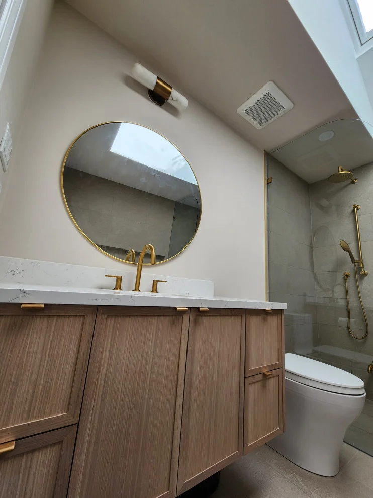 Light oak vanity with round gold mirror, gold faucet and toilet under sloped skylit ceiling